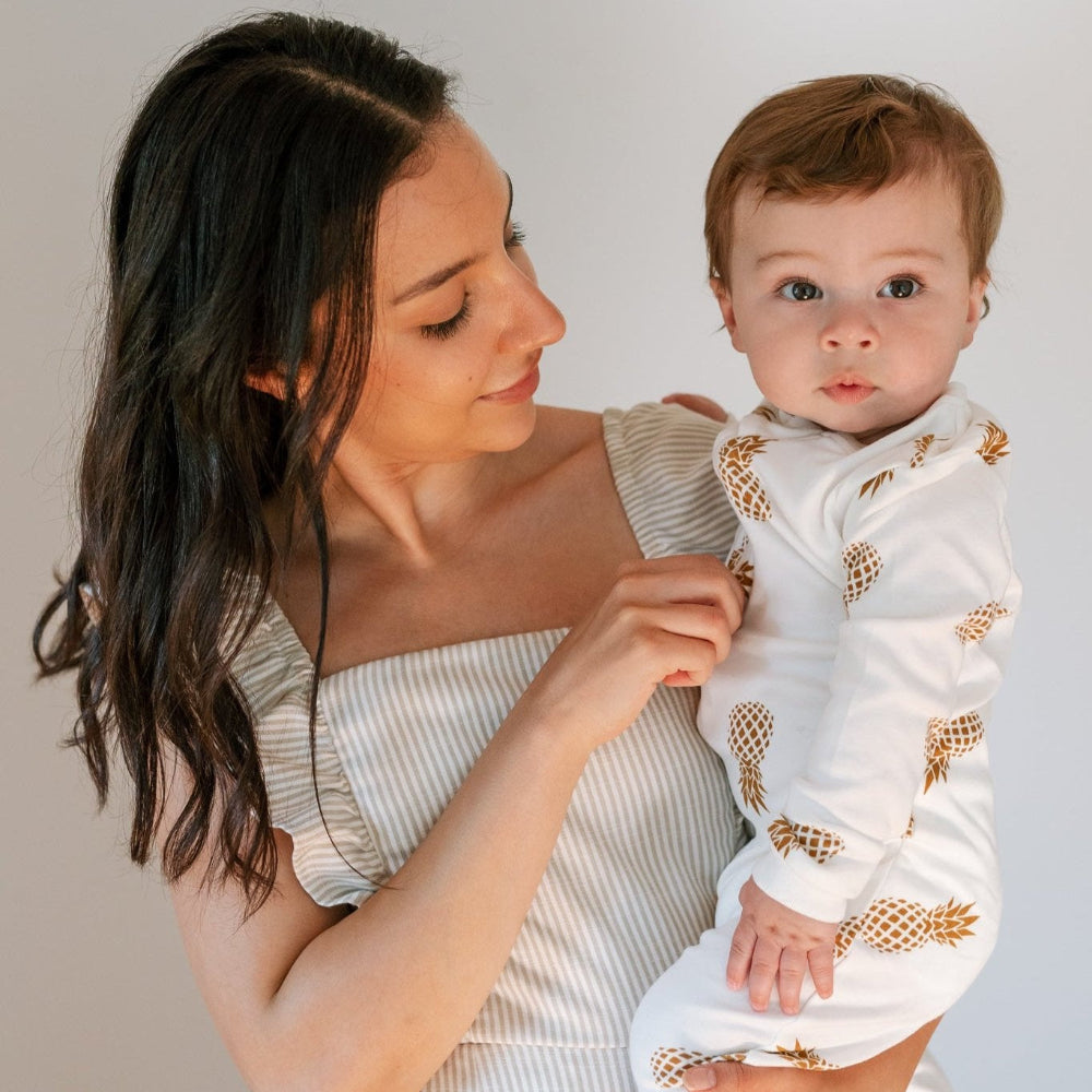 photo of a mother with dark hair and a striped dress holding a baby wearing ZippyJamz Sweet on the Inside baby sleeper - white background with gold pineapples