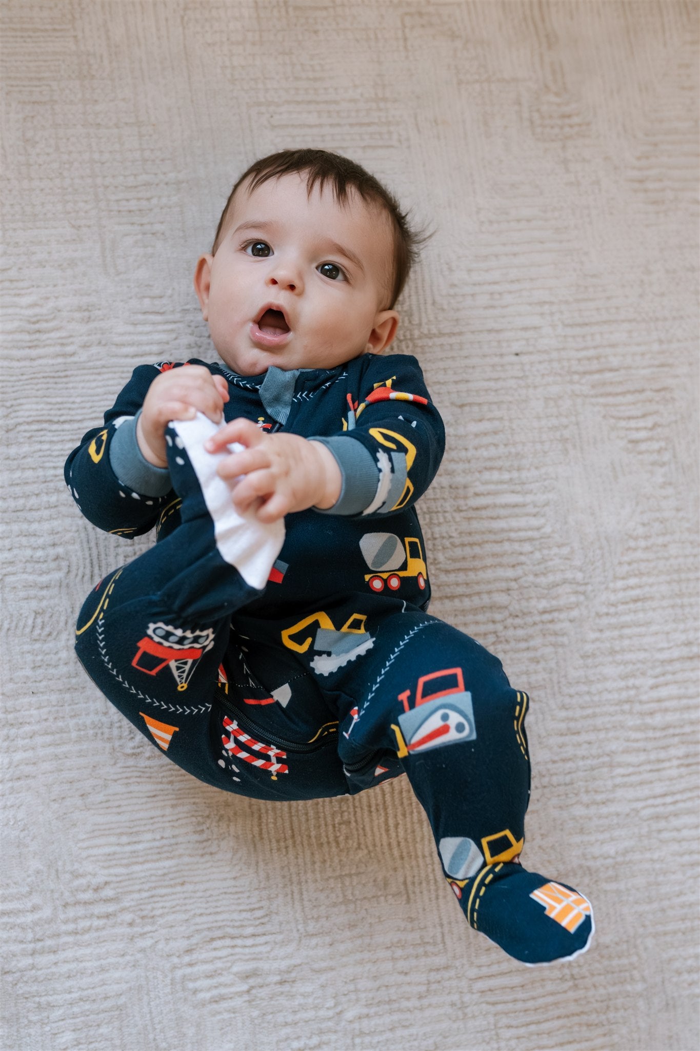 Baby lying on the ground holding its right foot while wearing a navy blue, construction patterned footed ZippyJamz sleeper