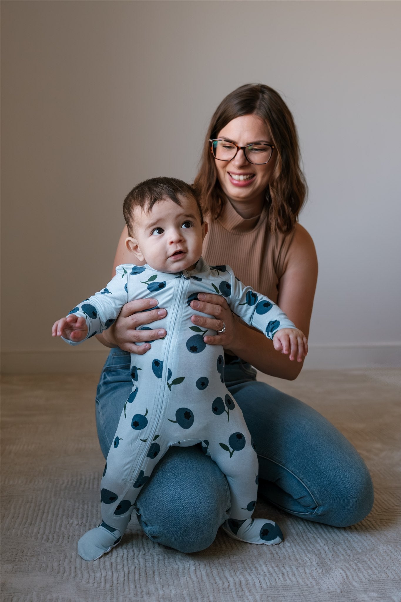 Adult holding a baby standing on a neutral carpet while wearing a blueberry patterned ZippyJamz footed baby sleeper