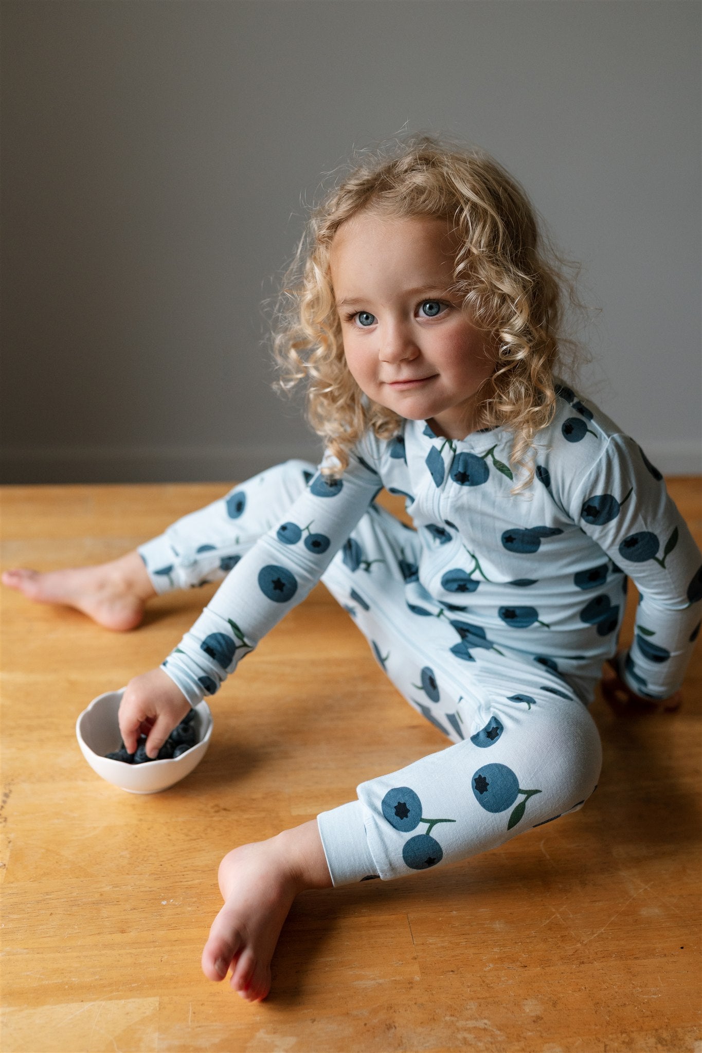 Blonde curly haired toddler sitting on a table with a bowl of blueberries wearing a footless blueberry patterned ZippyJamz sleeper