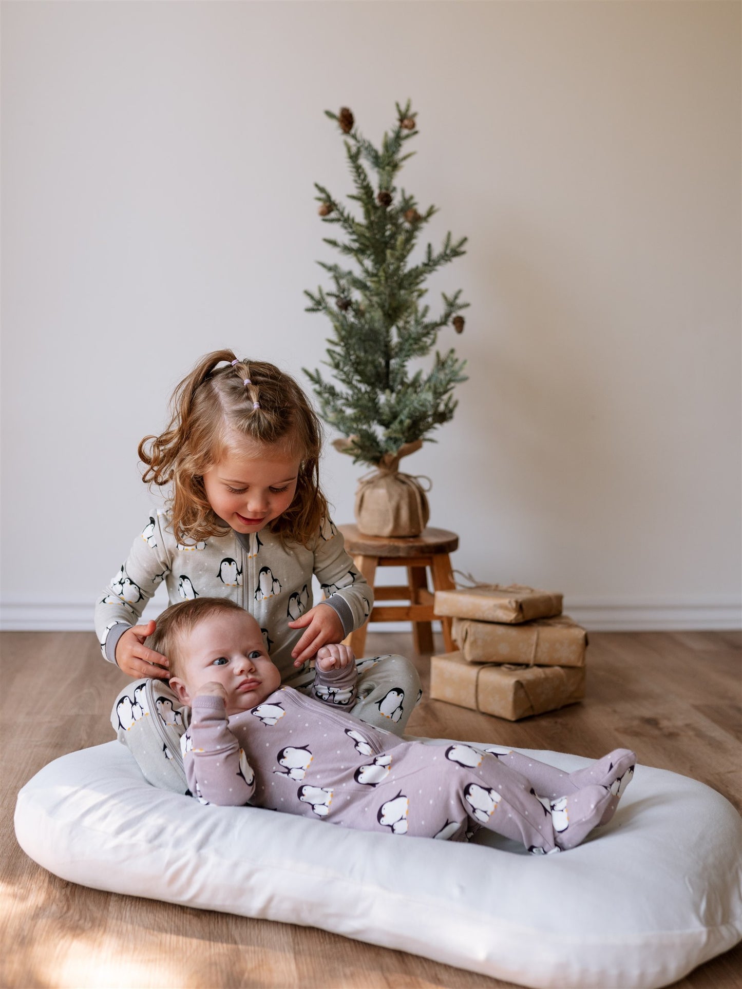 Two children in similar penguin baby pajamas sitting on a white cushion in front of a small Christmas tree with presents.