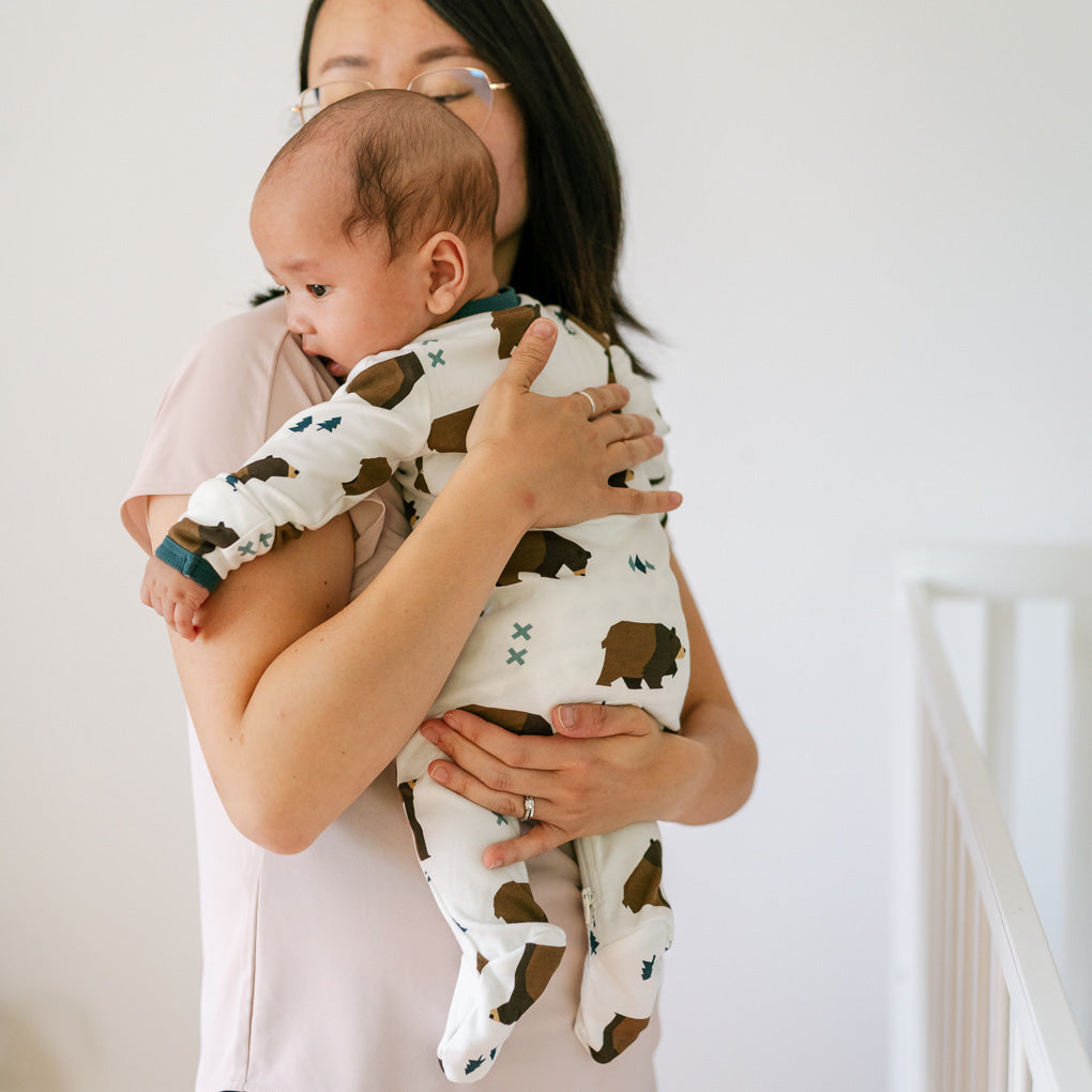 Woman holding a baby wearing ZippyJamz sleeper in Little Grizzle pattern with brown bears against a neutral backgroun
