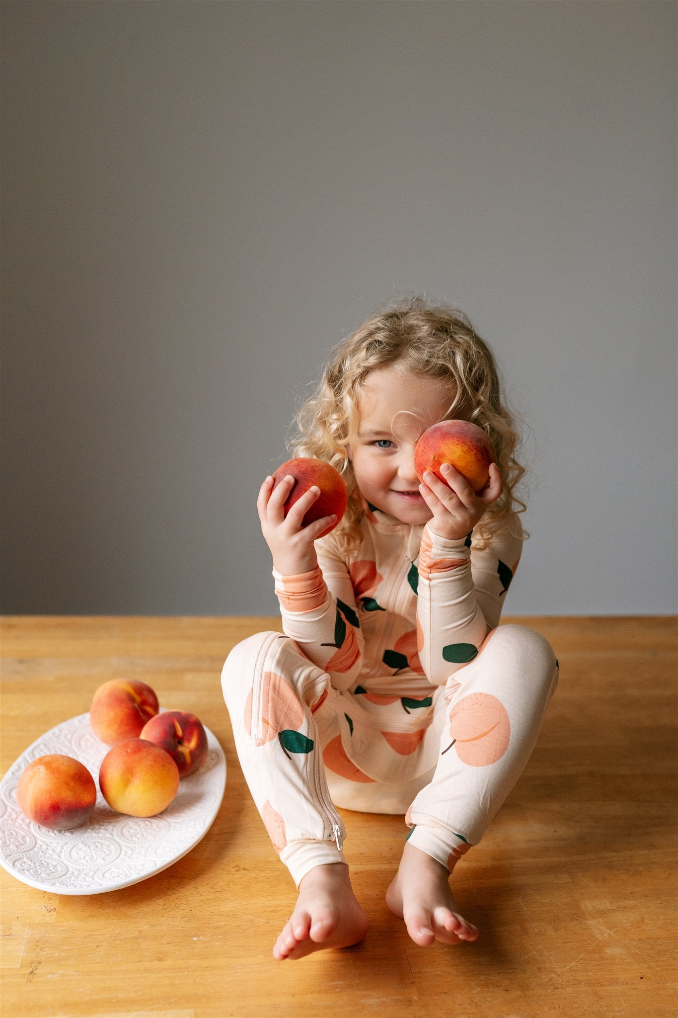 Child holding two peaches while wearing a footless peach patterned ZippyJamz sleeper sitting beside  a plate of peaches on a wooden table