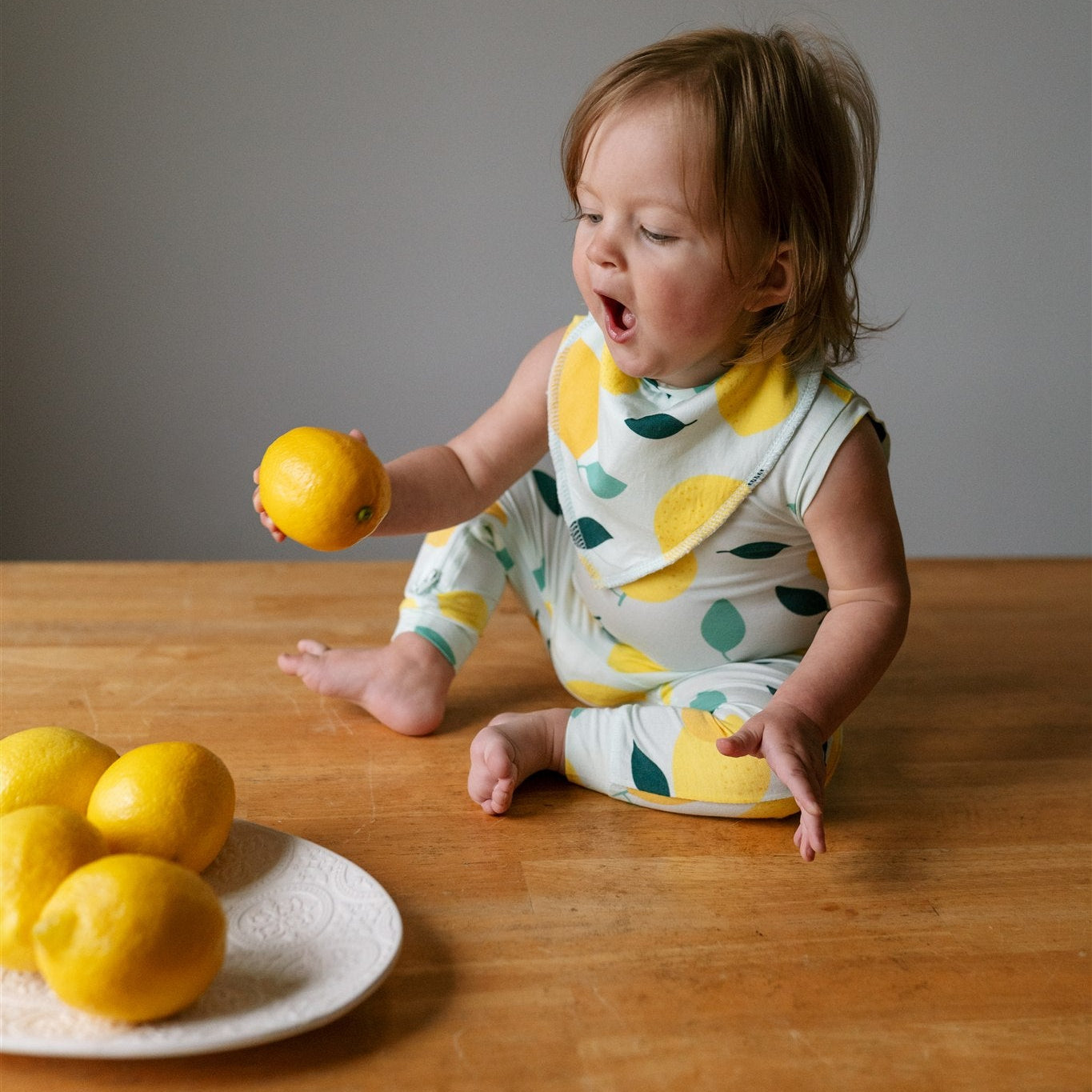 Baby in a lemon patterened ZippyJamz romper looking with open mouth surprise at a real lemon