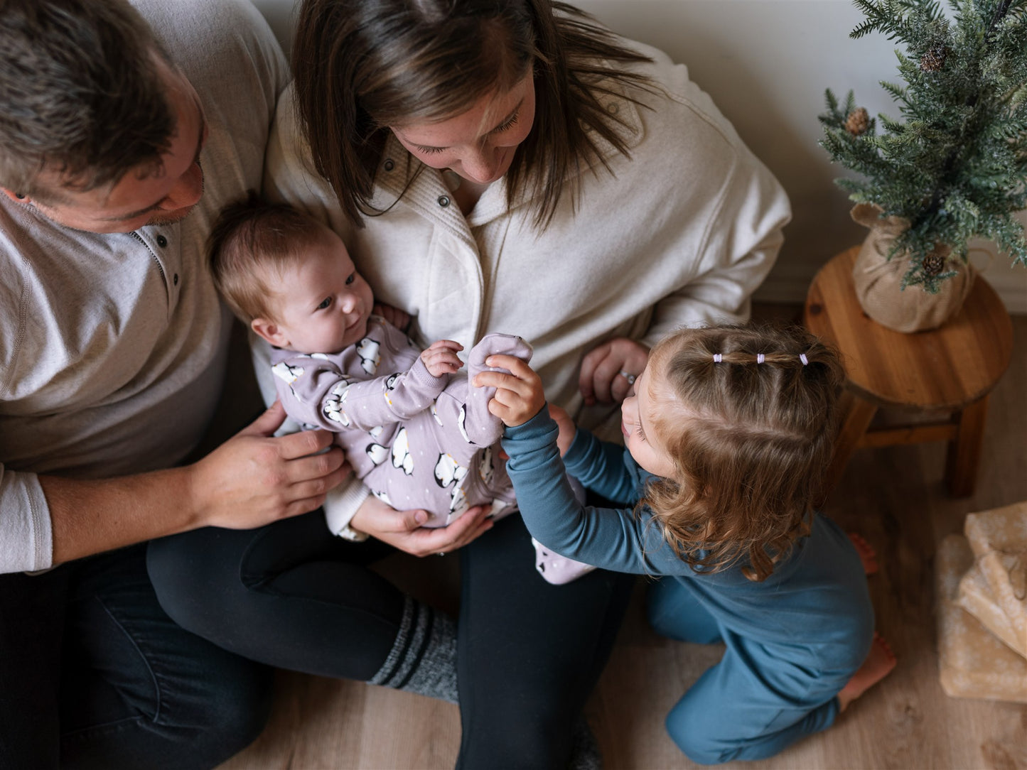 Family of four indoors, with a baby held by an adult,wearing a penguin baby sleeper and a toddler reaching out wearing a blue footless ZippyJamz sleeper