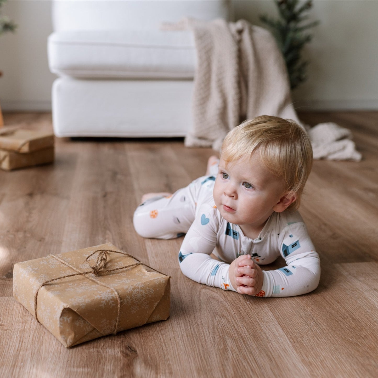 child lying on a wooden floor wearing ZippyJamz baby sleeper in Winter Whimsy pattern beside a wrapped gift