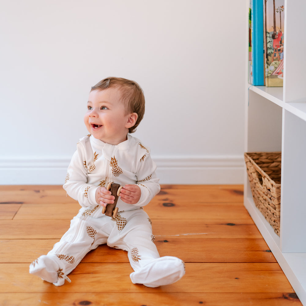 
                  
                    baby wearing white ZippyJamz sleeper with gold pineapple images sitting on a wooden floor smiling and holding a wooden toy car beside a white shelf
                  
                