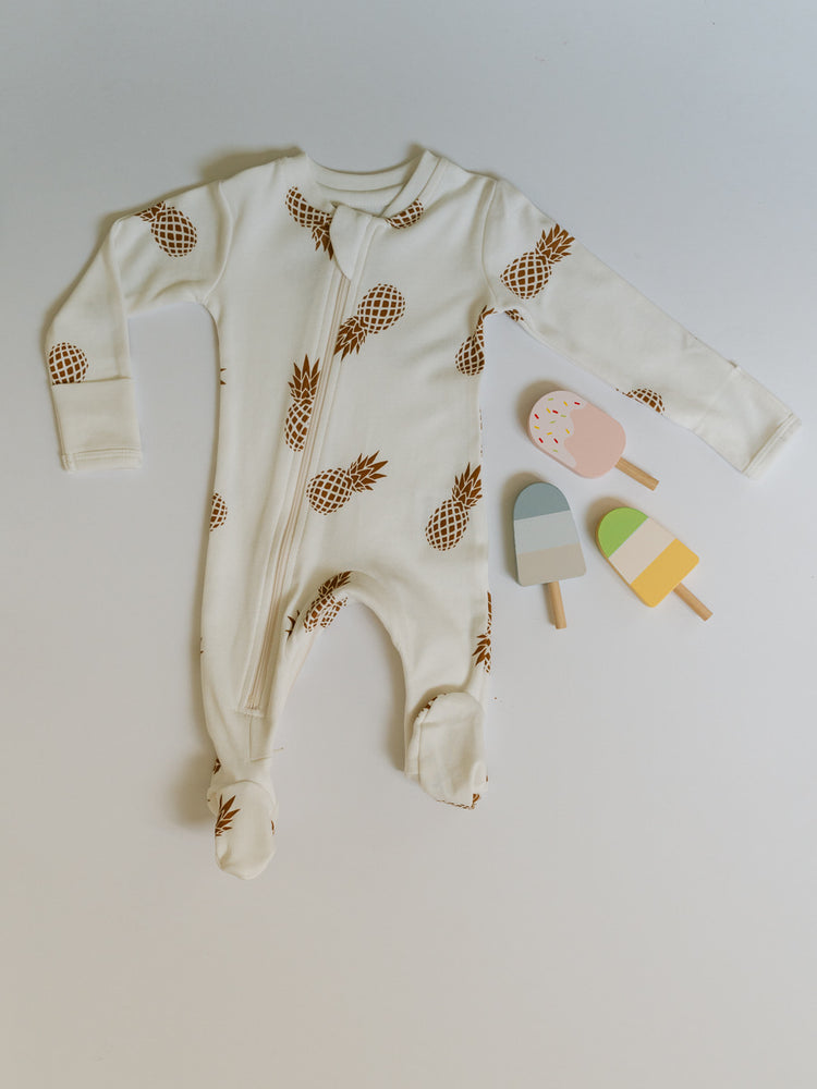 
                  
                    flat lay photo of ZippyJamz Sweet on the Inside baby sleeper - a white pajama with gold pineapple imagery showing the foldover cuffs available on newborn and 0-3M sizes. Photo includes toy popsicles 
                  
                